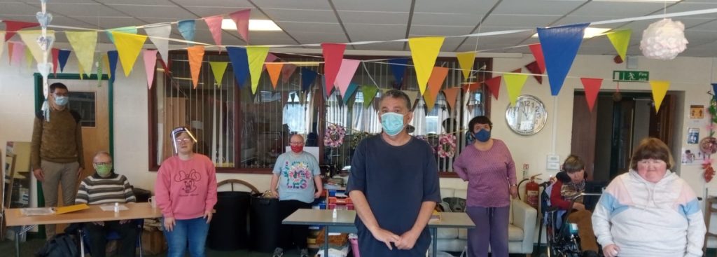 Image shows a group of adults with learning disabilities in a room standing at a social distance. They are wearing face masks or face shields to comply with current Covid precautions.